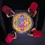 Vier Mönche in roten Roben gestalten ein farbenfrohes Sandmandala auf einer schwarzen Fläche, aufgenommen von oben. Schalen mit farbigem Sand stehen in der Nähe, während sie sorgfältig an dem komplizierten kreisförmigen Muster arbeiten.