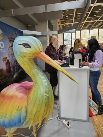 Eine große, farbenfrohe Reiherskulptur steht neben einem Stand, an dem zwei Frauen mit einer Besucherin in einem lila Pullover bei einer Indoor-Veranstaltung sprechen. Auf der Theke liegen Plakate und Broschüren, und im Hintergrund sind Menschen zu sehen.