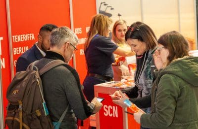 Eine Gruppe von Personen interagiert an einem Stand mit roten Wänden, die Reiseziele anzeigen. Zwei Mitarbeiter stehen hinter dem Stand, während drei Besucher mit Broschüren und einem Telefon hantieren.