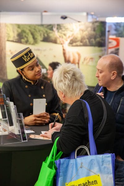 Eine Frau in einer Uniform und einem Hut mit der Aufschrift Victoria spricht mit zwei Personen an einem Stand. Die eine Frau schreibt, während sie eine grün-blaue Einkaufstasche hält. Im Hintergrund ist ein Display mit einem Hirschbild zu sehen.