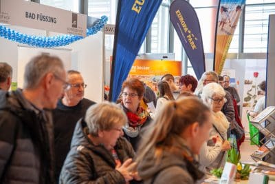 Eine Gruppe von Menschen interagiert und durchstöbert Informationsstände auf einer belebten Indoor-Ausstellung oder -Messe, mit Bannern und Displays im Hintergrund.