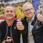 Zwei lächelnde Männer posieren zusammen, beide zeigen den Daumen nach oben. Sie tragen Jacken und große dekorative Halsketten und stehen vor einem bunten, verschwommenen Hintergrund.