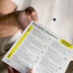 Eine Person in einem weißen Hemd hält eine gedruckte Aussteller-Checkliste mit geordneten Abschnitten und Aufzählungspunkten in deutscher Sprache für die Ausstellungsplanung und -vorbereitung.