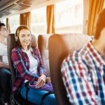 Eine junge Frau mit Kopfhörern und einem karierten Hemd sitzt lächelnd in einem Bus. Andere Fahrgäste sitzen in der Nähe. Das Sonnenlicht strömt durch die Fenster und schafft eine warme Atmosphäre.