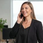 Eine Frau in einem schwarzen Blazer lächelt, während sie in einem modernen Büro mit unscharfen Schreibtischen und einer Pflanze im Hintergrund über ein Smartphone spricht.