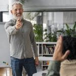Ein lächelnder Mann mit Bart, der eine Brille und ein graues Hemd trägt, steht und zeigt auf eine Person, die ihre Hand in einem Klassenzimmer oder Büro mit Bücherregalen und Pflanzen im Hintergrund hebt.