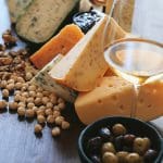 Verschiedene Käsescheiben mit sichtbaren Löchern und blauen Adern, umgeben von Walnüssen, Haselnüssen, einer Schale mit Oliven, einem Weinglas und einer kleinen Schale mit dunkler Marmelade auf einem Holztisch.