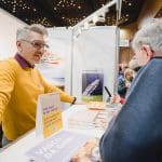 Ein Mann in einem gelben Pullover steht an einem Reisestand und spricht mit zwei Besuchern. Auf dem Tresen liegen Broschüren und ein Schild aus. Im Hintergrund sind Menschen unter dekorativen Deckenleuchten zu sehen.