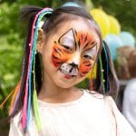 Ein lächelndes Kind mit bunten geflochtenen Haaren und Gesichtsbemalung in Form eines Tigers, das im Freien mit gelben und blauen Luftballons im Hintergrund steht.
