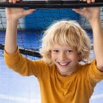 Lächelndes Kind mit gewelltem blondem Haar, das ein gelbes langärmeliges Hemd trägt und sich an einer schwarzen horizontalen Stange vor einem Netz festhält, mit einem blauen Hallensportplatz im Hintergrund.