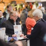 Ein älterer Mann, der eine Broschüre in der Hand hält, spricht mit einer Frau an einem Informationsschalter, umgeben von anderen Menschen in einem belebten Innenbereich. Beide scheinen in ein Gespräch vertieft zu sein, und auf dem Tresen liegt Informationsmaterial aus.