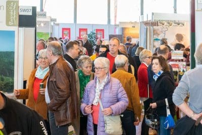 Eine große Gruppe von Menschen, meist ältere Erwachsene, geht durch eine belebte Veranstaltungs- oder Ausstellungshalle, in der im Hintergrund verschiedene Stände und Auslagen zu sehen sind.
