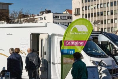 Menschen stehen in der Nähe eines weißen Wohnmobils, das neben städtischen Gebäuden geparkt ist. Auf einer grünen Fahne steht Frankana Freiko Camping ist bunt. Das Wetter ist sonnig und klar.