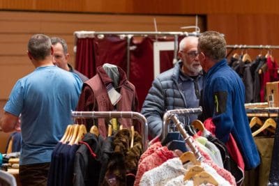 Mehrere Männer unterhalten sich und stöbern in den Regalen mit Jacken und Kleidern auf einem überdachten Markt oder Kleiderverkauf. Die Umgebung wirkt zwanglos und warm, mit verschiedenen Kleidungsstücken, die zur Schau gestellt werden.