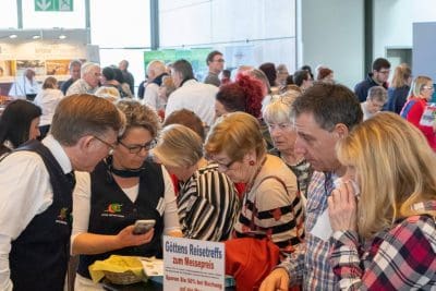 Eine große Gruppe von Menschen, viele mit grauen Haaren, versammelt sich bei einer Indoor-Veranstaltung um einen Stand, schaut sich Informationen genau an und spricht mit den Ausstellern. Ein Schild in deutscher Sprache bietet Reiseangebote an.