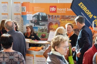 Besucher einer Reisemesse, die sich um einen Stand von Horst Becker Touristik mit Broschüren, Plakaten und Bannern versammelt haben; einige tragen orangefarbene Schals, und rechts ist eine Europa-Park-Flagge zu sehen.