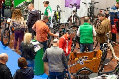 Eine Gruppe von Menschen stöbert auf einer Indoor-Fahrradausstellung in Fahrrädern und E-Bikes. Einige schauen sich die Fahrräder genau an, während andere sich unterhalten oder herumlaufen. Fahrräder und Fahrradzubehör sind auf dem Boden ausgestellt.