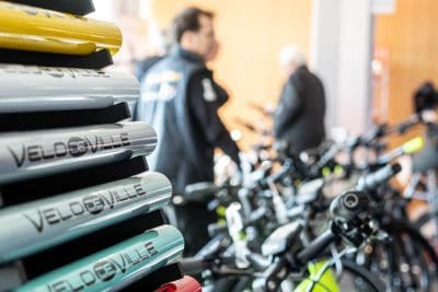 Nahaufnahme von gestapelten VELDEBILLE-Fahrradrahmen in verschiedenen Farben mit unscharfen Menschen und zusammengebauten Fahrrädern im Hintergrund, was auf ein Fahrradgeschäft oder eine Messe hindeutet.
