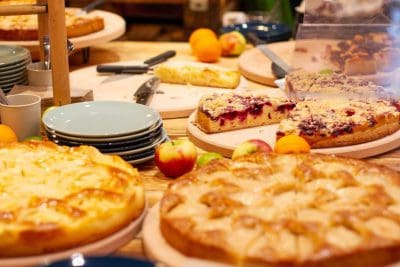 Ein mit verschiedenen Obstkuchen und -torten gefüllter Tisch, umgeben von frischen Äpfeln, Orangen, Stapeln von Tellern und Servierutensilien. Die Desserts sind teilweise aufgeschnitten und bereit zum Servieren.