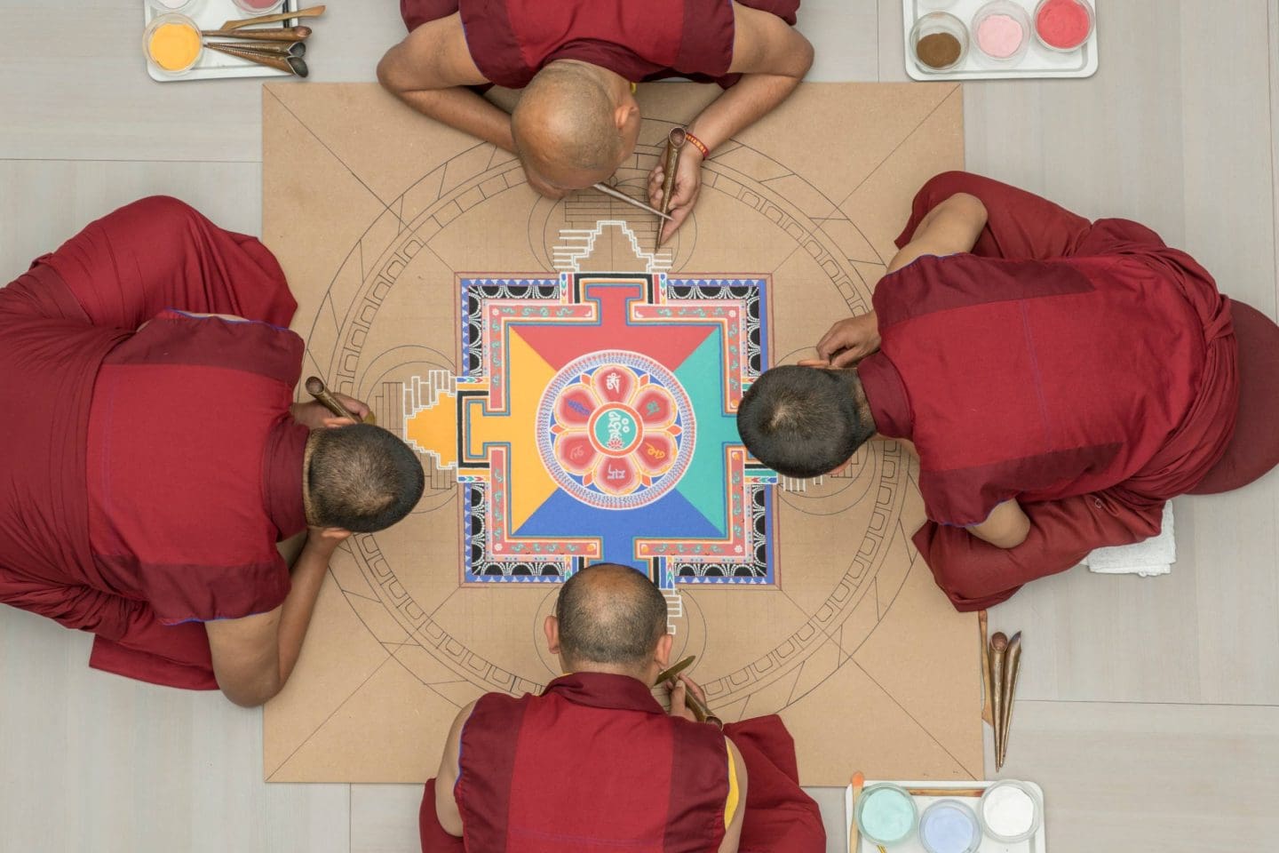 Vier Mönche in roten Roben erstellen ein farbenfrohes Sandmandala auf einer hellbraunen Fläche. Sie arbeiten mit feinen Werkzeugen und Farbschalen, von oben gesehen.