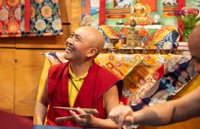Ein lächelnder buddhistischer Mönch in roter und gelber Robe hält hölzerne Instrumente in der Hand und sitzt vor farbenfrohen, verschnörkelten Dekorationen und einer goldenen Buddha-Statue. Im Vordergrund ist der Arm einer anderen Person zu sehen.