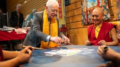 Ein Mann im Anzug und mit gelbem Schal hilft tibetischen Mönchen, ein buntes Sandmandala auf einem Tisch zu gestalten, während ein Mönch lächelt. Die Szene findet in einem Innenraum statt, mit lebhaften Dekorationen im Hintergrund.