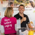 Ein lächelnder Mann zeigt hinter einem Tresen den Daumen nach oben, während eine Frau in einem rosa Hemd mit dem Aufdruck "Live Relax Enjoy" auf dem Rücken mit ihm spricht. Im Hintergrund sind Werbeplakate für Wellness zu sehen.