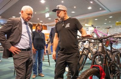 Ein Mann im Anzug hört einem anderen Mann in Freizeitkleidung zu, der bei einer Indoor-Veranstaltung die Funktionen eines Fahrrads erklärt. Im Hintergrund sind zwei weitere Personen und mehrere Fahrräder zu sehen.