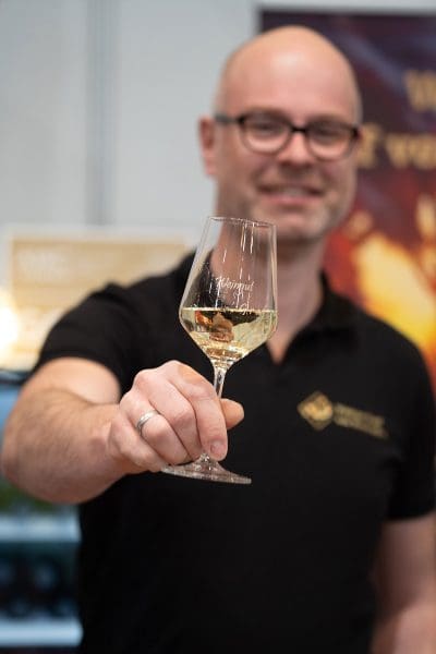 Ein lächelnder Mann mit Brille und schwarzem Hemd hält ein Glas Weißwein in die Kamera, der Hintergrund ist unscharf.