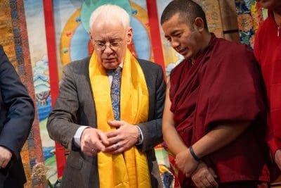 Ein älterer Mann im Anzug mit gelbem Schal spricht mit einem buddhistischen Mönch in kastanienbraunen Gewändern; im Hintergrund hängen bunte traditionelle Wandteppiche.