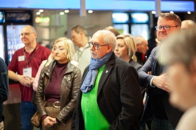 Eine Gruppe Erwachsener, sowohl Männer als auch Frauen, steht in einem Haus und hört aufmerksam zu. Im Mittelpunkt steht ein älterer Mann mit einer Brille, einem grünen Pullover und einem blauen Schal. Im Hintergrund sind helle Lichter und unscharfe Menschen zu sehen.