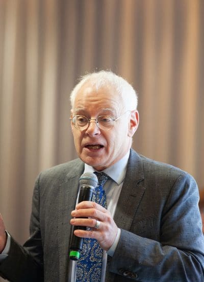 Ein älterer Mann mit Brille und weißem Haar, der einen Anzug und eine gemusterte blaue Krawatte trägt, spricht in ein Mikrofon. Der Hintergrund ist mit vertikalen Linien verschwommen.