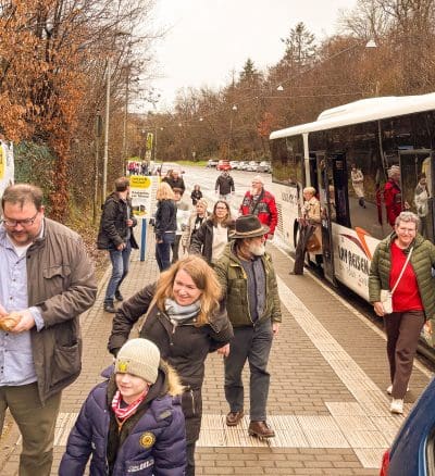 Menschen unterschiedlichen Alters gehen an einem bewölkten Tag in der Nähe einer Bushaltestelle spazieren; einige steigen aus einem weißen Bus aus, während andere den Bürgersteig entlang gehen. Bäume säumen den Hintergrund und die Szene wirkt lässig und lebendig.