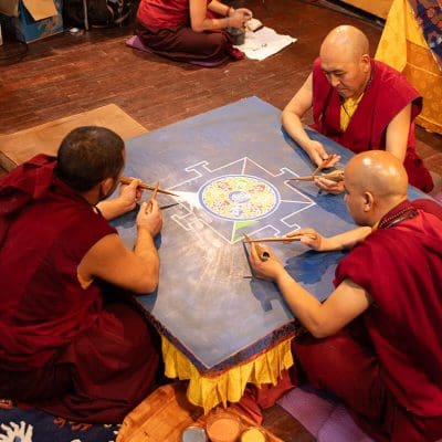 Drei Mönche in roten Roben sitzen um einen Tisch und gestalten sorgfältig ein buntes Sandmandala. Der Tisch ist mit blauem Tuch bedeckt, und in der Mitte entstehen verschlungene Muster. In der Nähe stehen Schalen mit farbigem Sand.