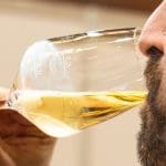 Nahaufnahme einer bärtigen Person, die ein Glas mit hellem, schaumigem Bier trinkt, das zum Mund hin gekippt ist. An der Seite des Glases sind Messmarkierungen sichtbar.